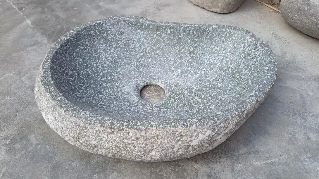 Un elegante lavatorio de piedra de río pulido instalado sobre un mueble de madera, mostrando la textura natural y el diseño de un ovalín de piedra moderno.
